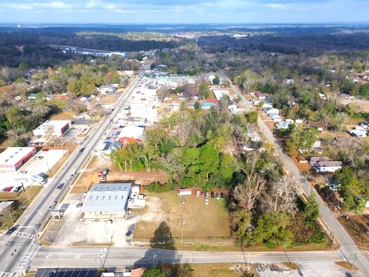 Oblique drone view over Dublin showing the commercial corridor of shops and service buildings with the wooded corner lot among them.